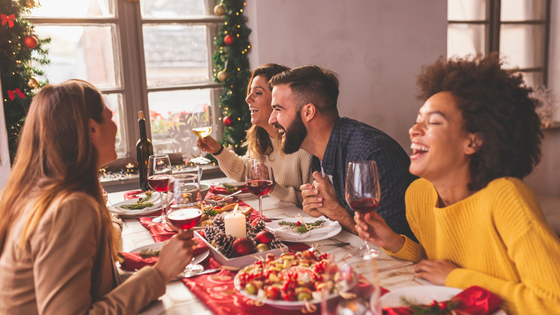 Group of people laughing and holding glasses of wine, seated around a table with holiday food and decorations.