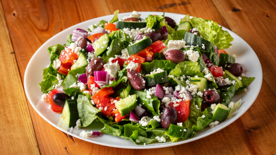 White bowl filled with chopped lettuce, cucumbers, red onions, tomatoes, olives, and feta cheese. 