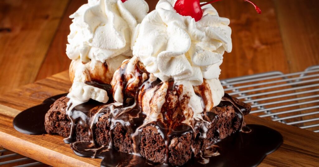 A large brownie covered in hot fudge, vanilla ice cream, and whipped cream with a cherry on a wooden board.