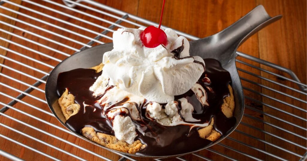 A pan filled with a baked chocolate chip cookie, covered in hot fudge and whipped cream with a cherry on top. 