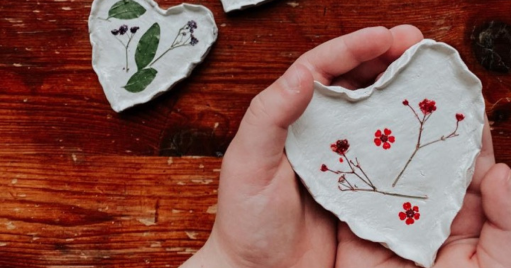 Hands holding a clay heart with pressed flowers on it. 